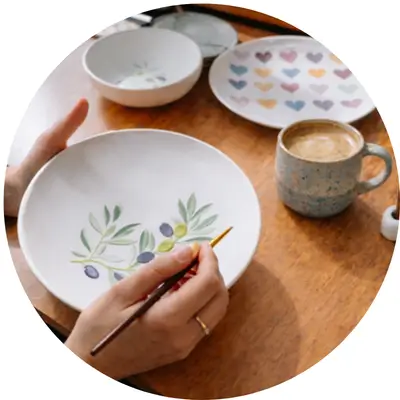 Keramikteller wird mit Blumenmuster bemalt bei gemütlicher Workshop-Atmosphäre mit Kaffeetasse